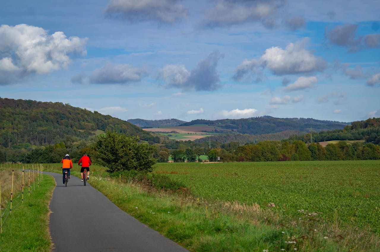 Two cyclists enjoying a ride on a rural path, surrounded by lush green fields and distant hills under a blue sky.